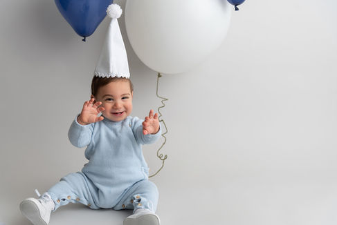 Little boy dressed in blue with a white party hat holding blue and white balloons