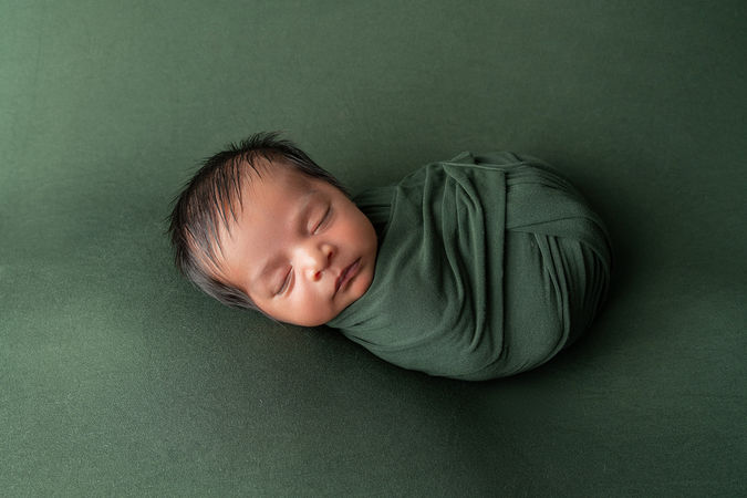 baby boy swaddled in dark green sleeps against dark green backdrop