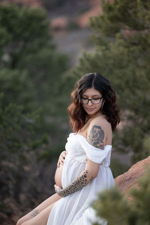 pregnant woman in white maternity gown cradles belly while sitting on a boulder