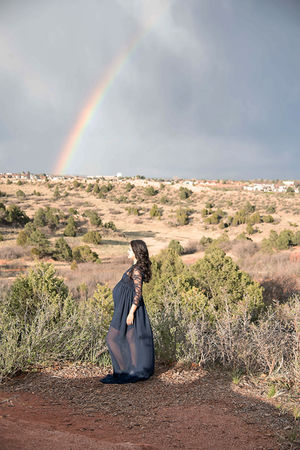 pregnant woman in black dress poses in front of rainbow