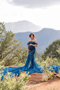 pregnant woman in blue dress cradles belly with mountains in the background