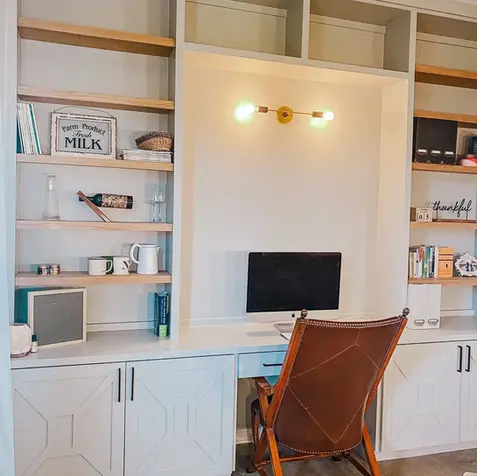 Home office with built-in shelves, desktop computer, a brown leather chair, and wall light fixture—ideal for your home renovation.
