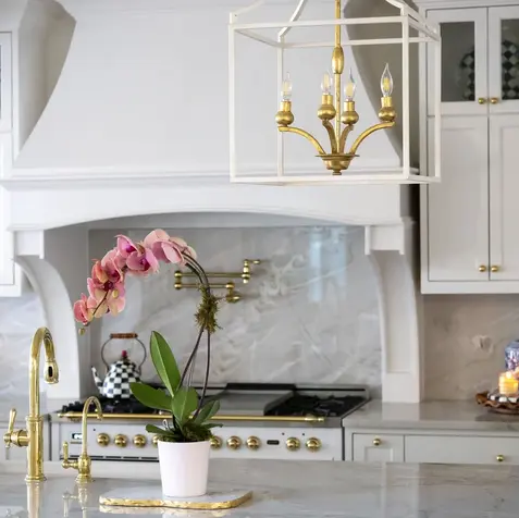 A modern kitchen with custom white cabinets, a marble countertop, a gold faucet, a potted orchid, and a white and gold chandelier.