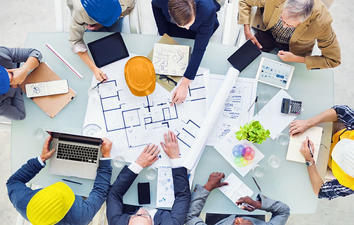 A group of professionals gathered around a table with architectural plans, various electronic devices, and work materials. The focus is on collaboration and planning in a professional setting