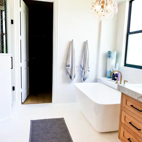 Modern bathroom with freestanding tub, wooden vanity, custom cabinets, dark floor mat, and geometric chandelier. Towels hang on wall.