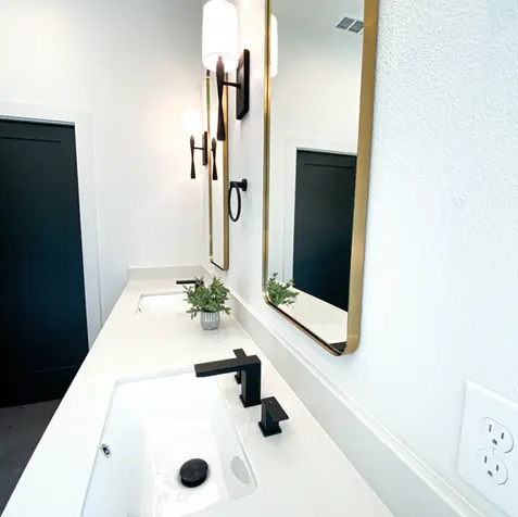 Modern bathroom with custom cabinets, a white countertop, black faucet, rectangular mirror, wall sconce, and small plant.