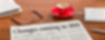 A wooden desk with a newspaper headline reading “Changes coming in 2019,” a pair of glasses lying on top, a red coffee cup on a saucer, and a white keyboard to the right. A pen is also placed beside the newspaper, and there’s an open newspaper in the background