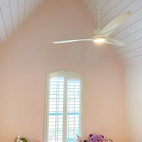 A pink-walled room with a vaulted ceiling, perfect for post-renovation relaxation, features a fan, window shutters, and cozy stuffed toys.