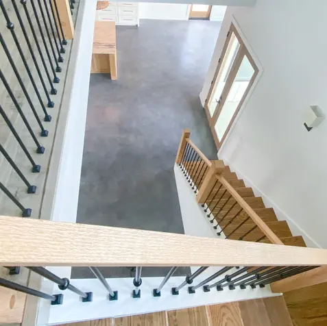 A view from the top of a wooden staircase with metal railings, looking down to a gray floor and dining table in the custom kitchen.