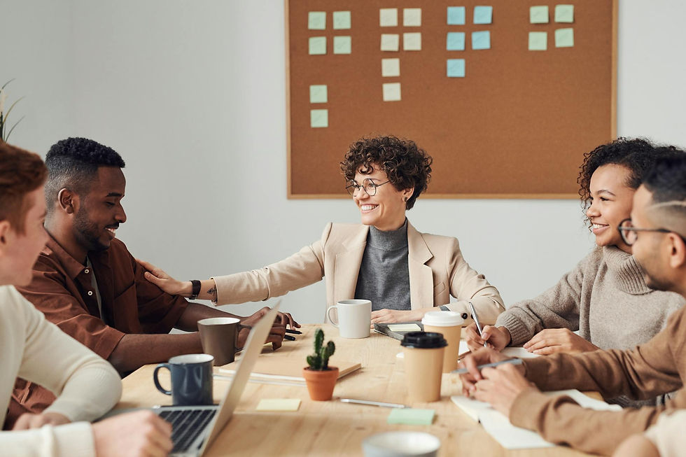 Diverse team of professionals engaged in a friendly discussion around a conference table, with a woman in a beige blazer smiling and placing a supportive hand on a colleague’s shoulder.