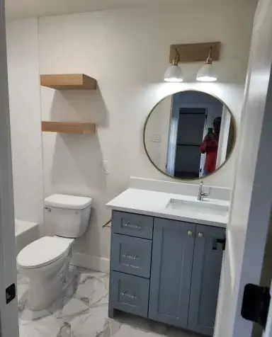 Bathroom remodel with a round mirror, gray vanity, white countertop, two wall shelves, and patterned floor tiles.