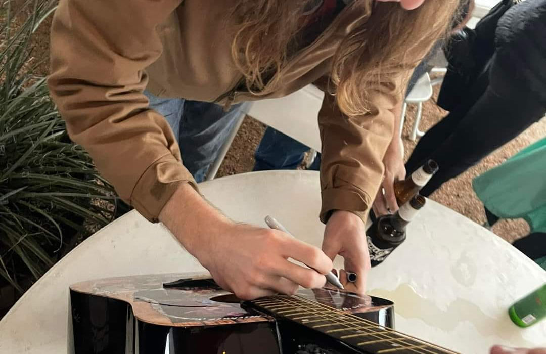Man signing a guitar on a table, writing on the fretboard detail.