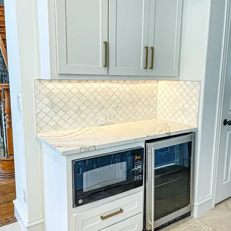 A kitchen corner with white cabinets, tile backsplash, a microwave on a shelf, and a small stainless steel fridge after home renovations.