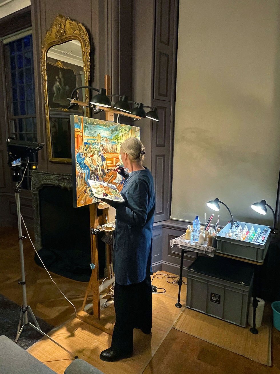 Eva schildert live aan een ezel in een statige kamer; verfpalet en lampen naast haar, warm licht en geconcentreerde sfeer.