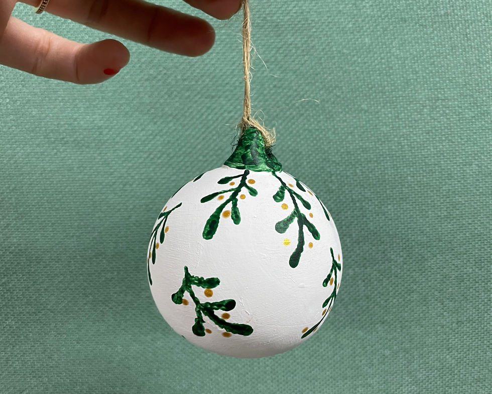 Hand holding a white painted ornament ball with green leaf sprigs and gold dots against a mint background.