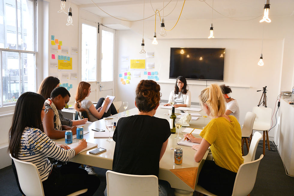 Diverse group in a bright meeting room takes notes around a table, with sticky notes on the wall and a large blank screen above.