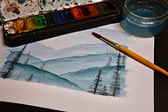 A watercolour brush, a paint pallette and a watercolour painting of mountains