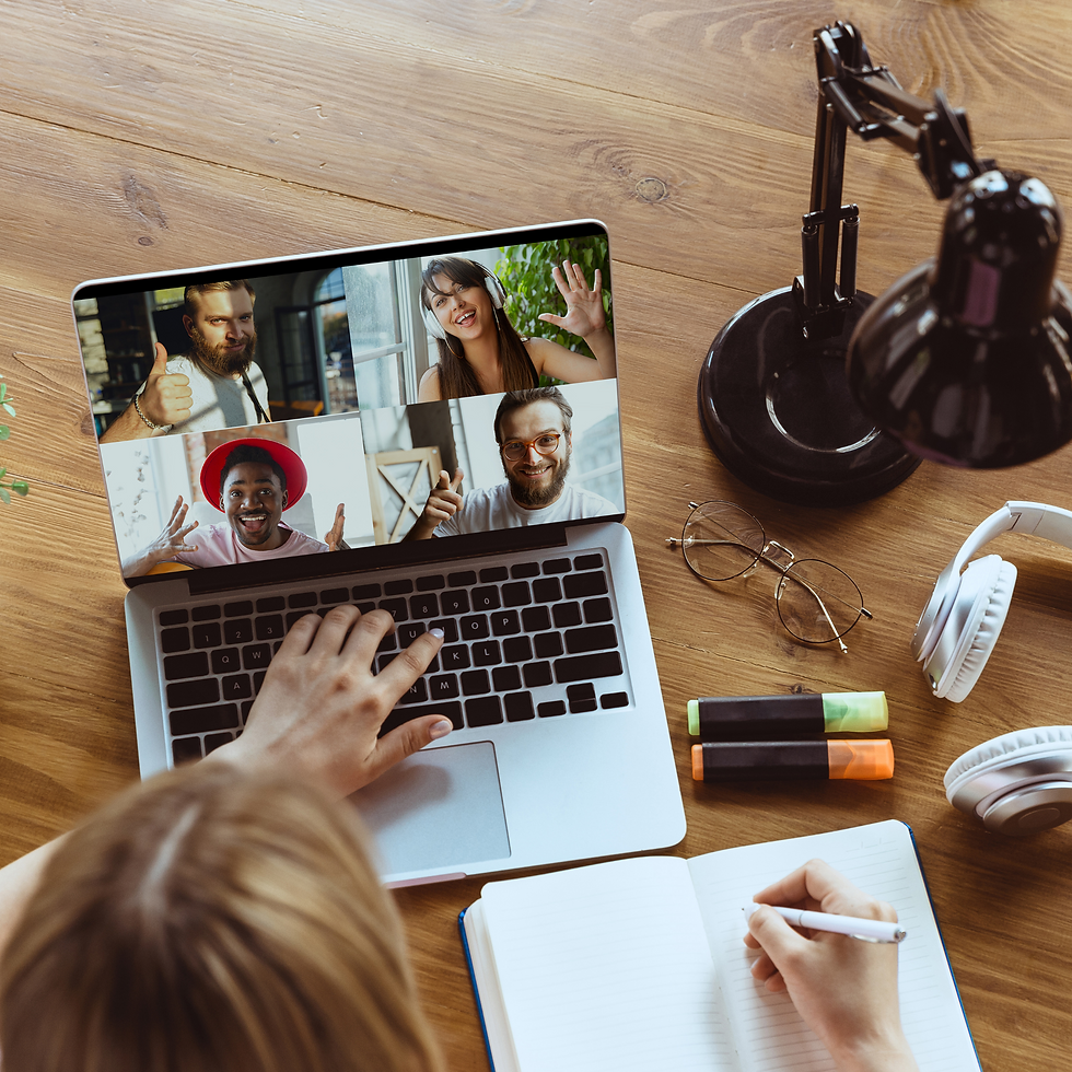 Person writing in notebook at desk while video calling four others on a laptop. Desk has headphones, glasses, markers, and lamp. Cozy mood.
