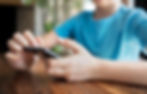 Child in a blue shirt using a smartphone, sitting at a wooden table. The background is blurred, suggesting an indoor setting.
