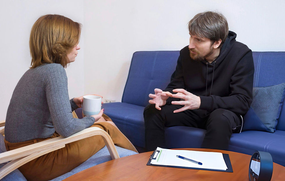 A woman holding a mug and a man talking are seated on a blue couch in a calm setting. A clipboard with a pen is on a table nearby.