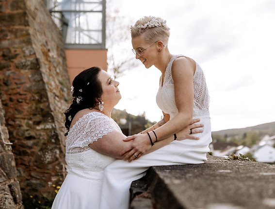 Lesbisches Paar auf Mauer in Burg bei Elopement in der Eifel. // Lesbian couple on a wall in a castle during elopment in Eifel (Germany).