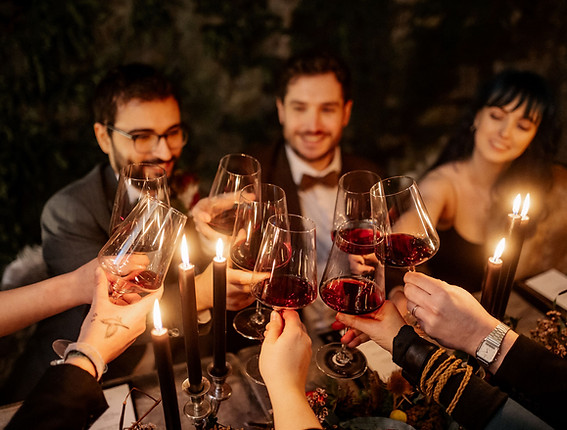 The wedding party at the grooms' table is clinking glasses with red wine to celebrate their dark Viking inspired wedding (gay wedding). Die Hochzeitsgäste am Tisch der Bräutigame stößt mit Rotwein an, um die dunkle Wikinger:innen-inspirierte Hochzeit zu feiern (schwule Hochzeit).