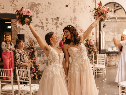 Queer wedding couple during their recessional at an industrial venue in Cologne (Germany). Their lesbian celebrant/officiant and guests are celebrating with them. Queeres Hochzeitspaar während Auszug in Industrie-Location (Harbour Club/New Yorker) in Köln (NRW bzw. Nordrhein-Westfalen). Ihre lesbische Traurednerin/Hochzeitsrednerin/freie Rednerin und Gäste feiern mit ihnen.