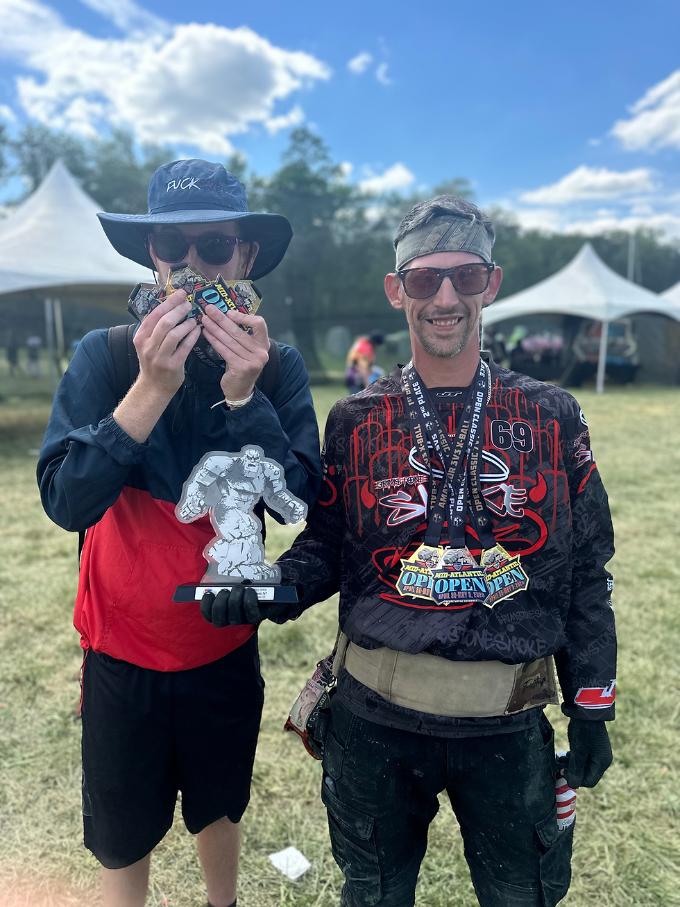 Two people outdoors, one holding a trophy and wearing medals; the other clutching Medals, wearing a hat. Both are smiling, tents in background.