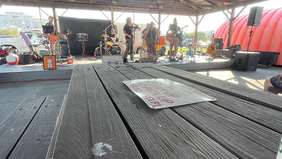 Band performs on an outdoor pavilion stage with guitars and drums; picnic table
