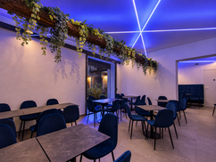 Sala con tavoli, sedie blu e trave decorata con piante e fiori sotto un soffitto illuminato di blu.