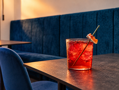 Cocktail rosso con ghiaccio su un tavolo scuro, davanti a una panca imbottita in velluto blu.