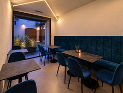 Sala con panca e sedie blu, tavoli scuri e un’ampia vetrata, illuminata da luci calde.