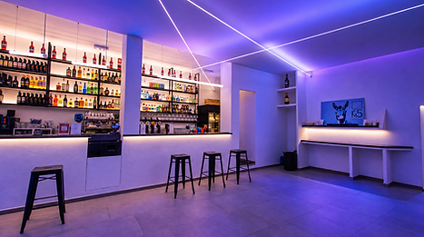 Bancone del cocktail bar KM5 a Torino con bottigliera illuminata e luci serali blu e viola.