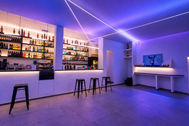 Veduta del bar con bancone chiaro, sgabelli neri, bottigliera illuminata e luci blu e viola al soffitto.