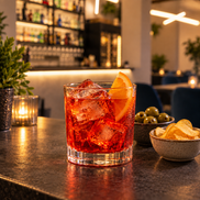Cocktail rosso con ghiaccio e fetta d’arancia sul bancone, accanto a piccole ciotole e una luce da tavolo.
