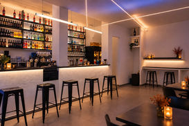 Ampio bancone con sgabelli neri, scaffali di bottiglie e sottili linee luminose che attraversano il soffitto.
