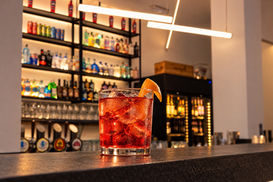 Cocktail rosso con ghiaccio e guarnizione d’arancia sul bancone, con bottigliera e frigorifero sullo sfondo.