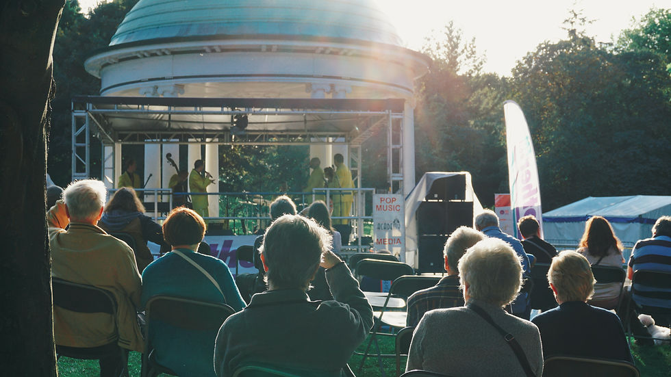 Publikum sitzt bei Open-Air-Konzert vor Bühne im Park; Musiker in Gelb, Schild mit POP MUSIC, MEDIA, warmes Sonnenlicht.