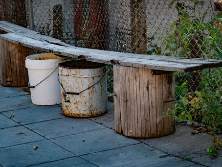 Rustic bench made of wood planks on buckets, beside a chain-link fence. Weathered look, surrounded by greenery on a stone path.