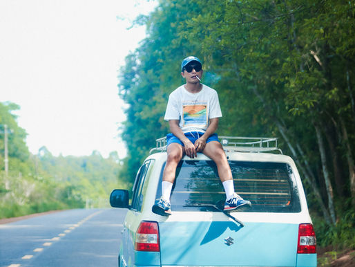 Man in sunglasses sits on a blue van, smoking, on a tree-lined rural road. A folding chair is in the foreground. Relaxed vibe.