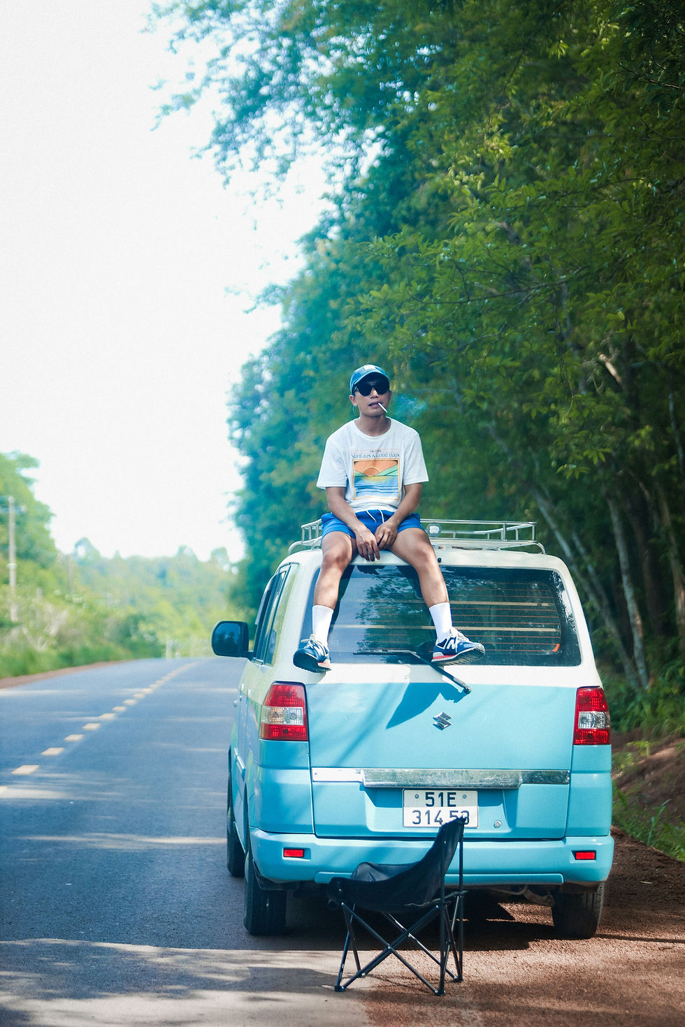Man in sunglasses sits on a blue van, smoking, on a tree-lined rural road. A folding chair is in the foreground. Relaxed vibe.