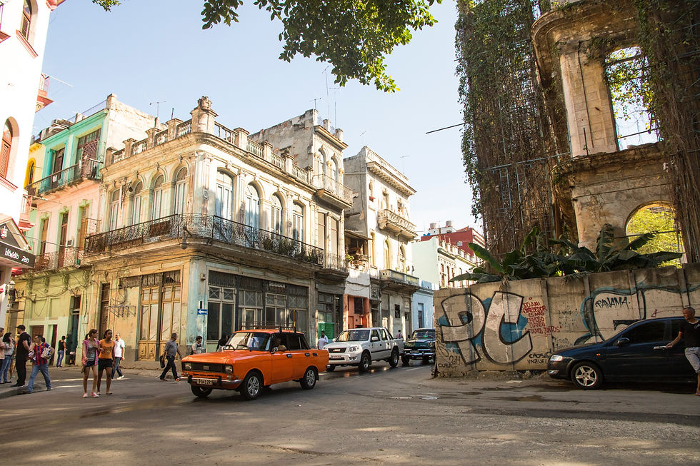 Carros em rua de cidade com prédios antigos ao fundo. Pessoas caminham. Grafite cobre muro à direita. Atmosfera animada e ensolarada.