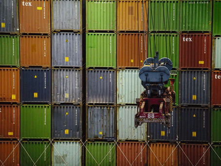 A crane lifts a container in front of colorful stacked shipping containers. "Tex" is visible on some containers. Industrial setting.