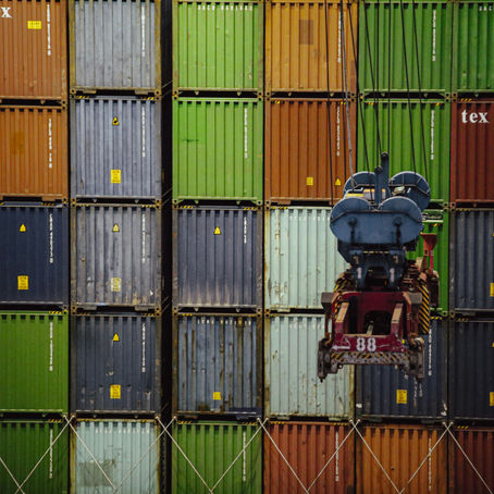 A crane lifts a container in front of colorful stacked shipping containers. "Tex" is visible on some containers. Industrial setting.