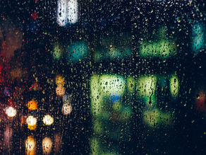 Raindrops on a window with blurry city lights in the background. Dark, moody atmosphere with reflections of colored lights.