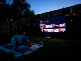 Couple on a blanket outside at night, watching a large TV screen with movie selections. Cozy ambiance with string lights and cushions.