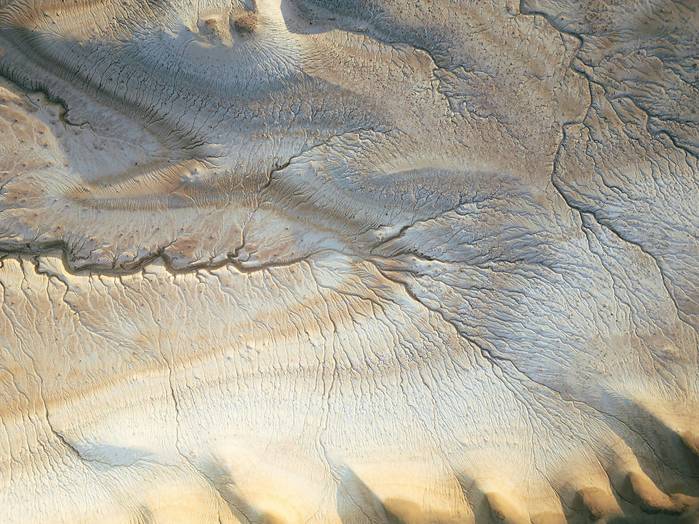 Aerial view of eroded beige desert hills with branching dry channels and rippled textures, like a weathered abstract landscape.