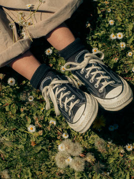 Legs in a beige skirt, black socks and sneakers lying in a field of tiny white daisies and dandelions.