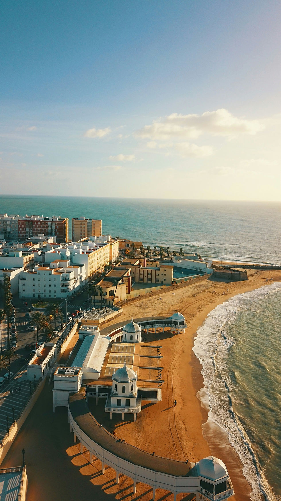 An aerial view of a picturesque coastal city, showcasing a blend of modern and traditional architecture. The golden sandy beach curves gently along the turquoise sea, bordered by a line of palm trees and colorful buildings under a bright, clear sky.