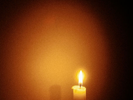 Lit candle in a dark room, casting warm yellow light and shadow on the wall. The mood is peaceful and serene.